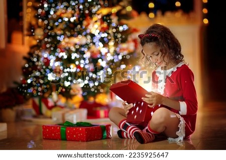 Child opening present at Christmas tree at home. Kid in Santa pajamas dress with Xmas gifts and toys. Little girl with gift box at fireplace. Family celebrating winter holidays. Home decoration.