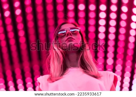 Teen hipster girl in stylish glasses standing on red tunnel with neon light wall background, female teenager fashion model pretty young woman looking at night club city light glow Royalty-Free Stock Photo #2235933617
