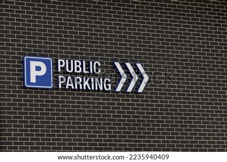 Parking road sign with arrows on a brick wall. "Public Parking" in text with directional arrows, as well as blue and white "P" parking sign mounted on modern style a dark colored brick wall 