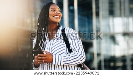 Business woman is looking away and smiling. She is standing in the city with a smartphone in her hand. Woman commutes to work in the morning. Royalty-Free Stock Photo #2235949091
