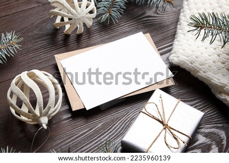 Christmas greeting card mockup with envelope, gift box, green fir tree branches and holiday decorations on brown wooden background. White New Year card with winter decor