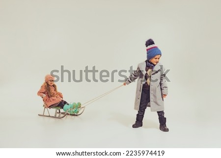 Portrait of boy and girl, children in winter clothes sledding isolated over grey studio background. Winter holidays. Concept of childhood, friendship, fun, lifestyle, fashion, retro style