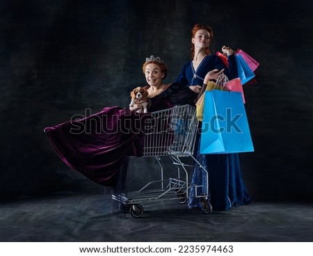 Portrait of two beautiful women, royal persons doing shopping isolated on dark background. Sitting on shopping trolley. Concept of comparison of eras, modernity and renaissance, baroque style, history