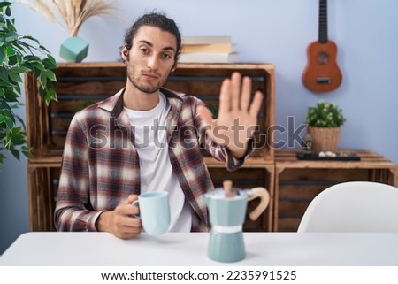 Young hispanic man drinking coffee from french coffee maker with open hand doing stop sign with serious and confident expression, defense gesture 