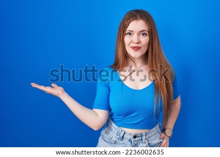 Redhead woman standing over blue background smiling cheerful presenting and pointing with palm of hand looking at the camera. 