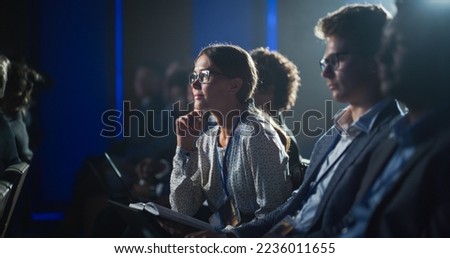 Young Woman Sitting in a Crowded Audience at a Business Conference. Female Attendee Writing Down Important Bullet Points into a Notebook. Keynote Speech in Auditorium with Successful Businesspeople. Royalty-Free Stock Photo #2236011655