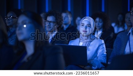 Arab Female Sitting in a Dark Crowded Auditorium at a Tech Conference. Young Muslim Woman Using Laptop Computer. Specialist in Hijab Watching Innovative Technology Presentation. Royalty-Free Stock Photo #2236011657