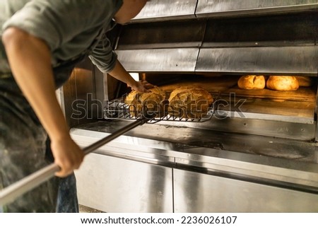 unrecognizable baker taking loaves out of the oven of the artisan bread factory Royalty-Free Stock Photo #2236026107