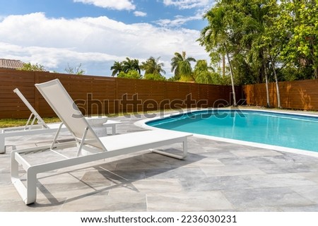 point of view of turquoise pool area with two sun loungers, wooden fence, palms and trees, blue sky Royalty-Free Stock Photo #2236030231