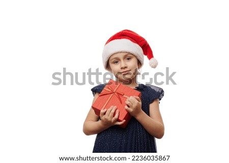 Caucasian beautiful smiling little girl dressed in Santa Claus hat and elegant evening dress, posing with cute gift box with Christmas present, white background. Happy festive mood of winter holidays