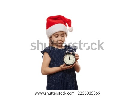 Beautiful little girl in Santa hat and evening dress, holding a black retro alarm clock showing midnight on dial, ready to celebrate the upcoming christmas or New Year, isolated on white background