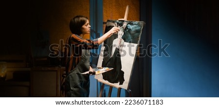 A young artistic girl, a painter is working on oil painting artwork. In her hands are brushes and a palette. A woman creates an art piece in the mysterious atmosphere of her studio, workshop at night.