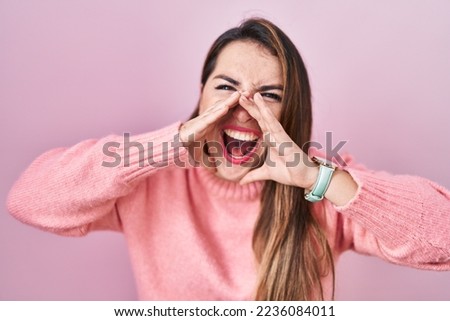 Young hispanic woman standing over pink background shouting angry out loud with hands over mouth 