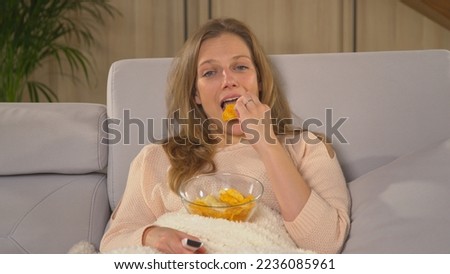 CLOSE UP: Relaxed woman eating snacks and watching entertaining TV program. Pretty lady covered with blanket and munching on chips while watching movie. Young woman enjoying on a winter evening.