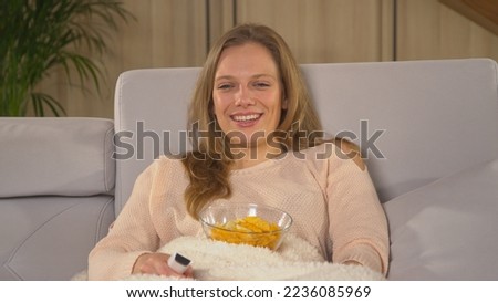 CLOSE UP: Beautiful woman enjoying on a comfy sofa and watching romantic comedy. Pretty lady covered with blanket and eating snacks while watching movie. Young woman relaxing on a winter evening.