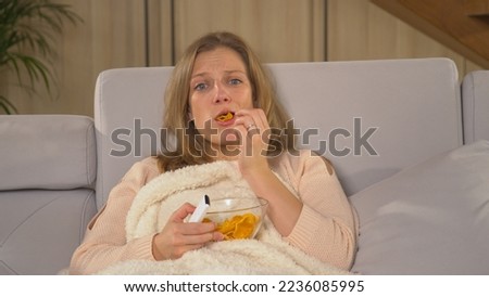 CLOSE UP: Pretty woman becoming stressed at watching unpleasant TV news report. Pretty woman covered with blanket and eating snacks while following TV news. Woman on a comfy sofa watching TV report.