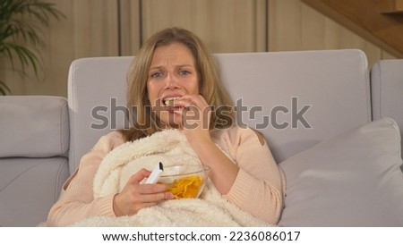 CLOSE UP: Young lady becoming upset while watching latest television news report. Pretty woman covered with blanket and eating snacks while following TV news. Woman on a comfy sofa watching TV report.
