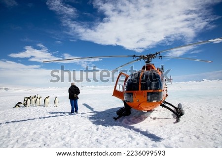 Helicopter landed on sea ice with pilot and emperor penguins in the background on a sunny day