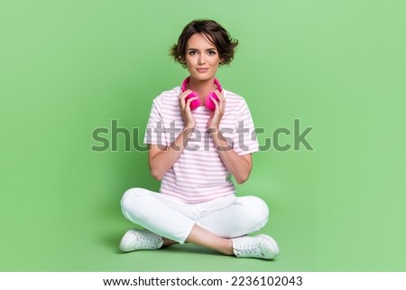 Full length photo of nice young woman crossed legs weekend touch earphones dressed trendy striped look isolated on green color background