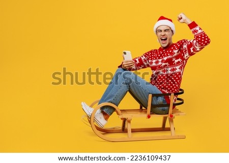 Full body fun merry young man wear red knitted Christmas sweater Santa hat posing sledding use mobile phone do winner gesture isolated on plain yellow background. Happy New Year 2023 holiday concept
