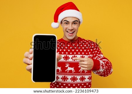Merry young man wear red knitted christmas sweater Santa hat posing hold show use mobile cell phone blank screen workspace area isolated on plain yellow background. Happy New Year 2023 holiday concept