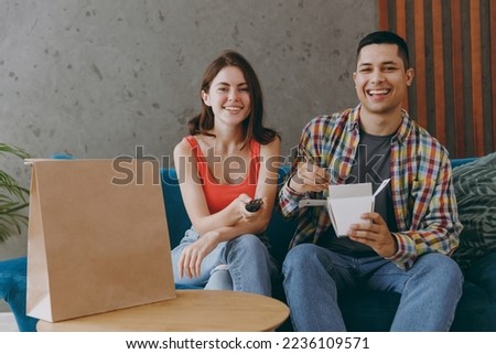 Young couple two friends man woman wear casual clothes eat Chinese food cuisine in takeaway carton container box sit on blue sofa together watch tv stay at home rest spend time in living room indoor