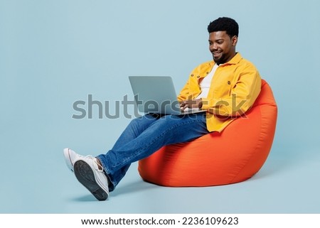 Full body smiling happy young man of African American ethnicity he wear yellow shirt sit in bag chair hold use work on laptop pc computer isolated on plain pastel light blue background studio portrait Royalty-Free Stock Photo #2236109623