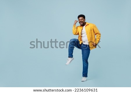 Full body excited young man of African American ethnicity 20s he wear yellow shirt doing winner gesture celebrate clenching fists say yes isolated on plain pastel light blue background studio portrait