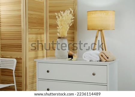 Vase with dried flowers, stacked towels and candles on chest of drawers in room. Interior design