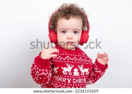 Beautiful little baby boy with curly hair wearing red Christmas knitted sweater against white background enjoys listening to videos or music wearing red headphones, clapping and dancing. 