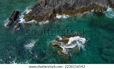 Black moss-covered rocks among turquoise waves of the Celtic Sea. White sea foam on the waves. Waters of the Atlantic Ocean. Aerial photo.