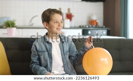 Smiling boy playing alone in the living room with an orange balloon prepared for his birthday party. A lonely, friendless, introverted, antisocial boy is looking at the right side of the screen.