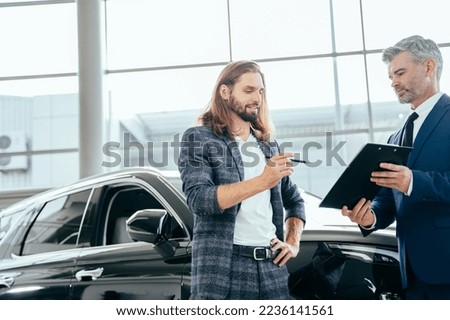 Successful middle-aged businessman is selling cars to male buyer in a car showroom. Handsome customer with car dealer agent making deal and signing on agreement document contract in car dealership.