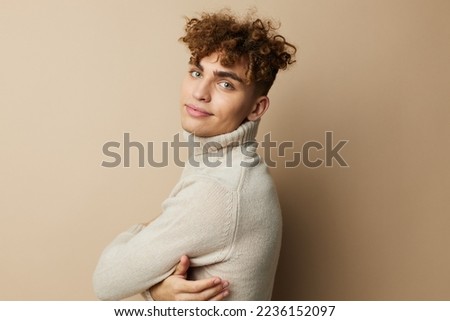 portrait of a handsome man with curly hair, standing on a beige background in a light turtleneck, holding his hand on his body, looking pleasantly into the camera.