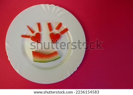 a smile with heart-shaped eyes is carved from a watermelon and laid out on a white round plate on a red background.  view from above