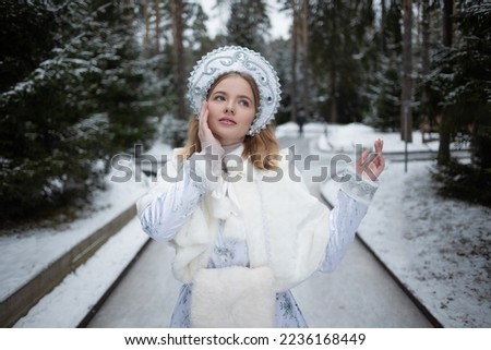 Snow Maiden , a cute young blonde in a white snow maiden costume with a kokoshnik , in the forest among snow - covered fir trees