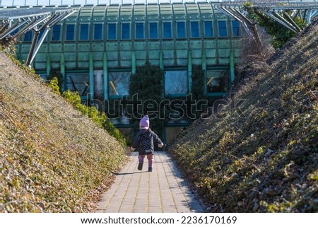 a little girl walks between two hills on the roof of Warsaw University Library outside in sunny weather, a child in a pink hat is wearing pink pants and a jacket, photo in early spring