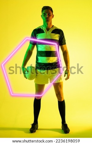 Serious male caucasian rugby player with ball by illuminated triangle standing on yellow background. Copy space, composite, sport, shape, competition, confident, playing, match and abstract concept.