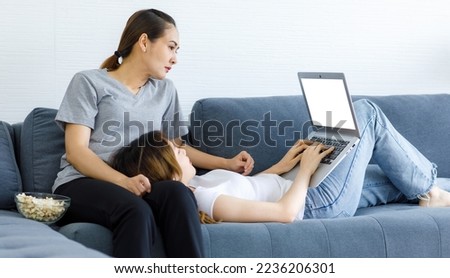 Millennial Asian young happy cheerful female lesbian laying down on LGBTQ lover girlfriend lap holding credit card using laptop computer browsing surfing internet shopping online at home living room.