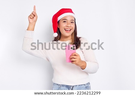 Smiling young Asian woman in a Christmas hat holding mobile phone and pointing finger up, creating genius solutions isolated over white background. celebration Christmas holiday and New Year concept