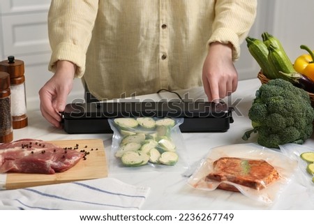 Woman using sealer for vacuum packing with plastic bag of zucchini at white table in kitchen, closeup Royalty-Free Stock Photo #2236267749