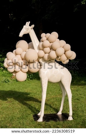 A white cream giraffe sculpture stands in the park. Exotic theme party decoration in nature, outside. Sculpture, large round, many balloons on the neck of a giraffe. Green and black natural background Royalty-Free Stock Photo #2236308519