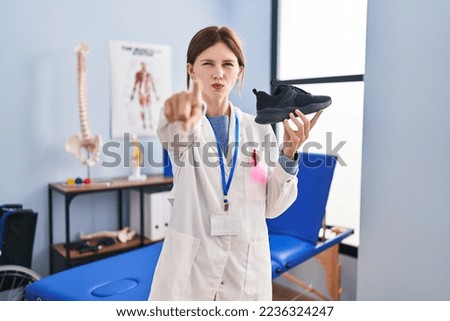 Young physiotherapist woman holding sneakers pointing with finger to the camera and to you, confident gesture looking serious 