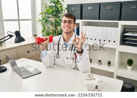 Young hispanic doctor man holding dumbbells doing ok sign with fingers, smiling friendly gesturing excellent symbol 