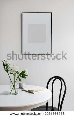 open book, glass vase with blossom flowers and ceramic cup on round wooden table, black chair, empty or template poster in dark frame on white wall in living room, vertical shot