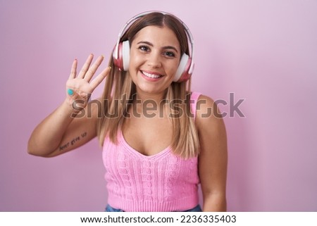 Young blonde woman listening to music using headphones showing and pointing up with fingers number four while smiling confident and happy. 