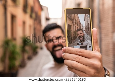 View of a screen with a young man with a beard wearing a turtleneck taking a selfie sticking out his tongue in one of the most prominent streets of his city.