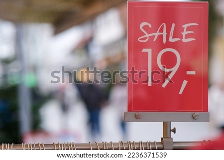 Close-up of a red price tag that says 'Sale' and the number '19' on a clothes rack outside a shop with people on the pedestrian street in the blurred background