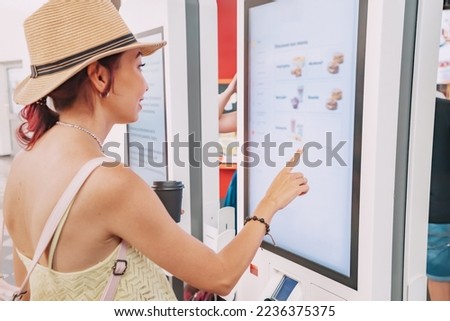 A female customer uses a touchscreen terminal or self-service kiosk to order at a fast food restaurant. Automated machine and electronic payment Royalty-Free Stock Photo #2236375375