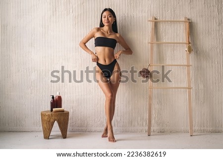 Full length shot of korean woman in bikini posing near textured wall in the bathroom